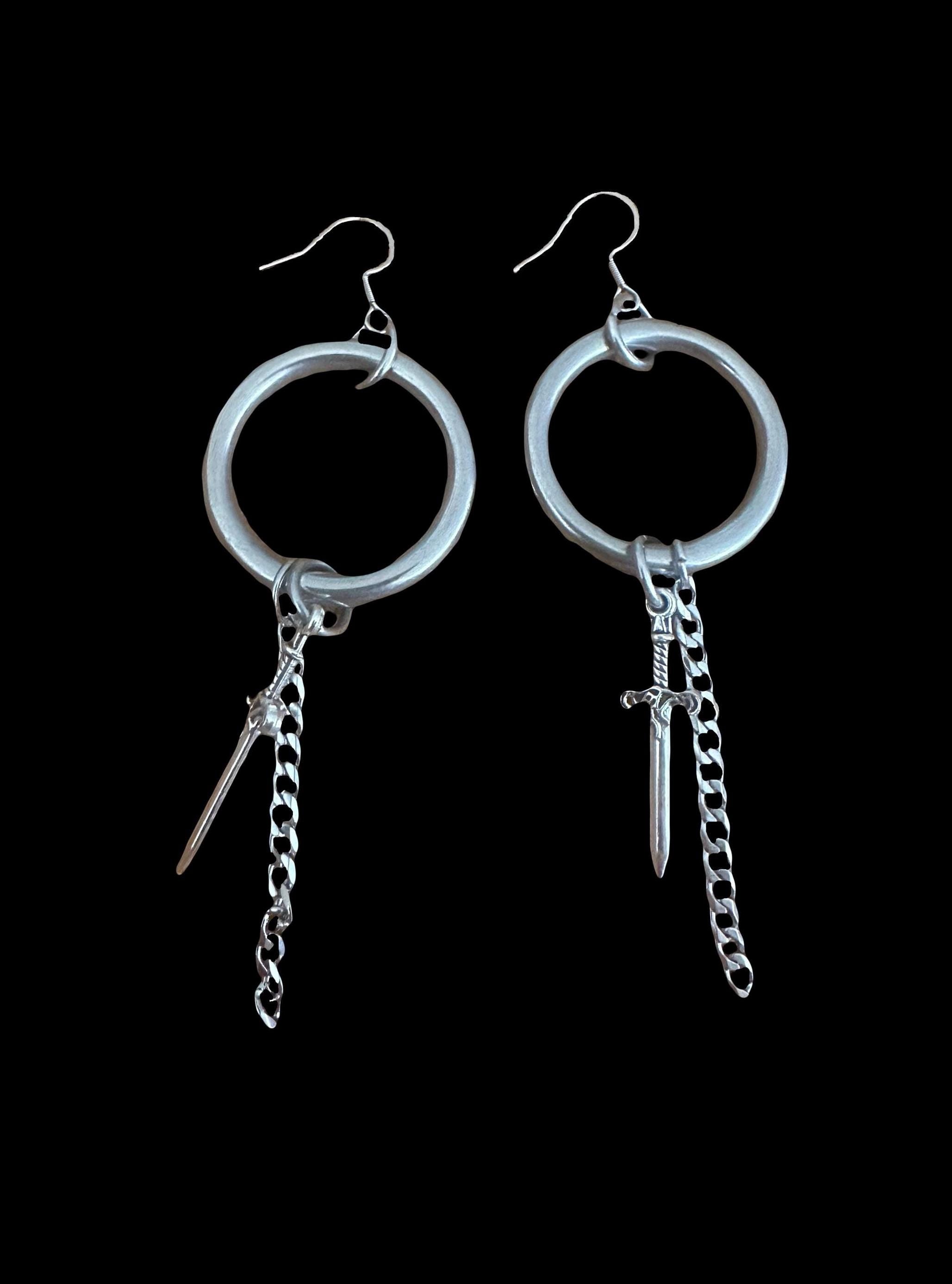 May include: Silver dangle earrings feature circular hoops with a dangling chain and sword charm.  The chain also includes a smaller, pointed charm. The earrings have a minimalist, edgy aesthetic, suitable for a modern or gothic style.