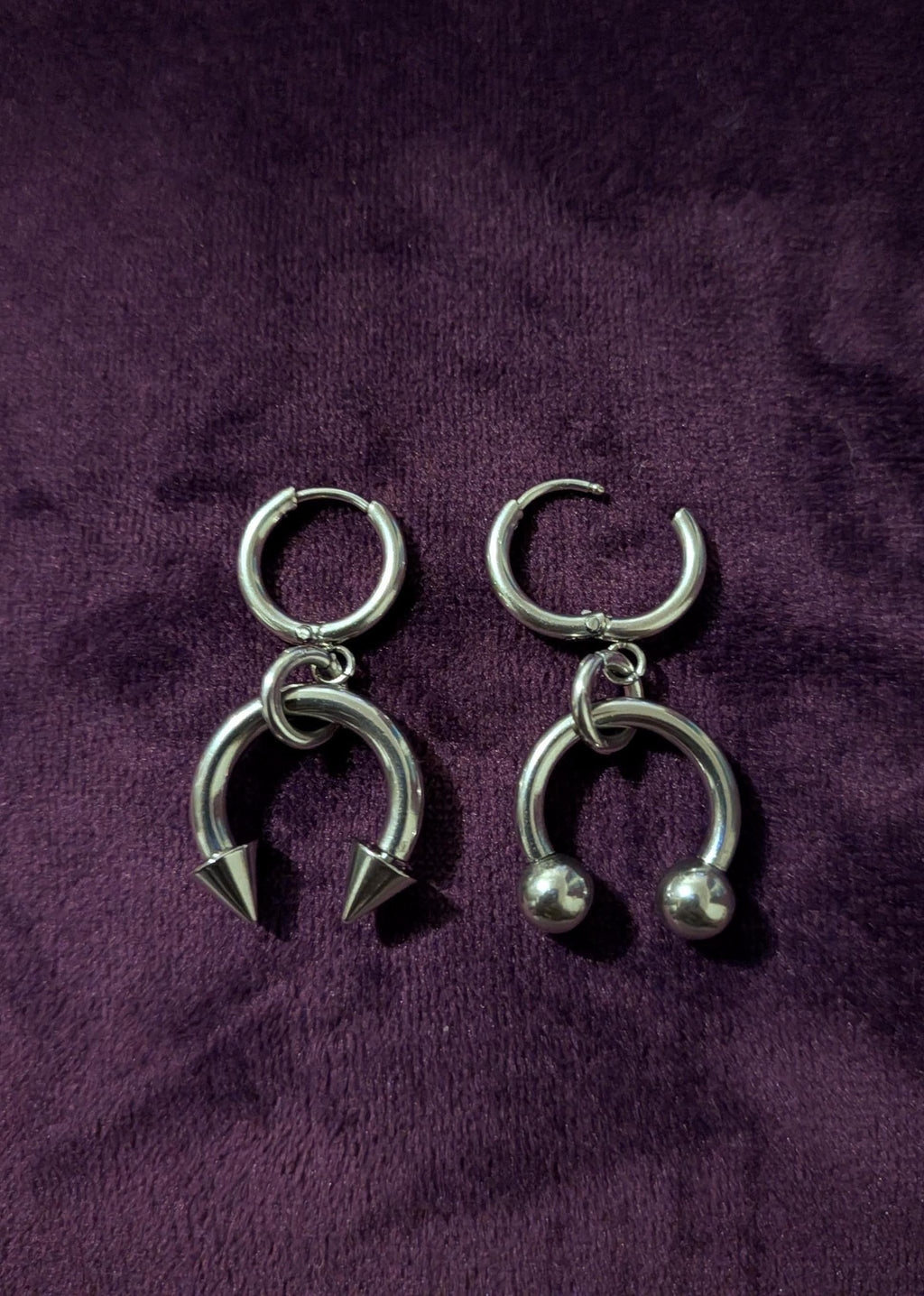 May include: A pair of silver-colored earrings. One earring features a hoop with a downward-facing, pointed spike. The other earring has a hoop with two small, round balls. Both earrings have a circular top.