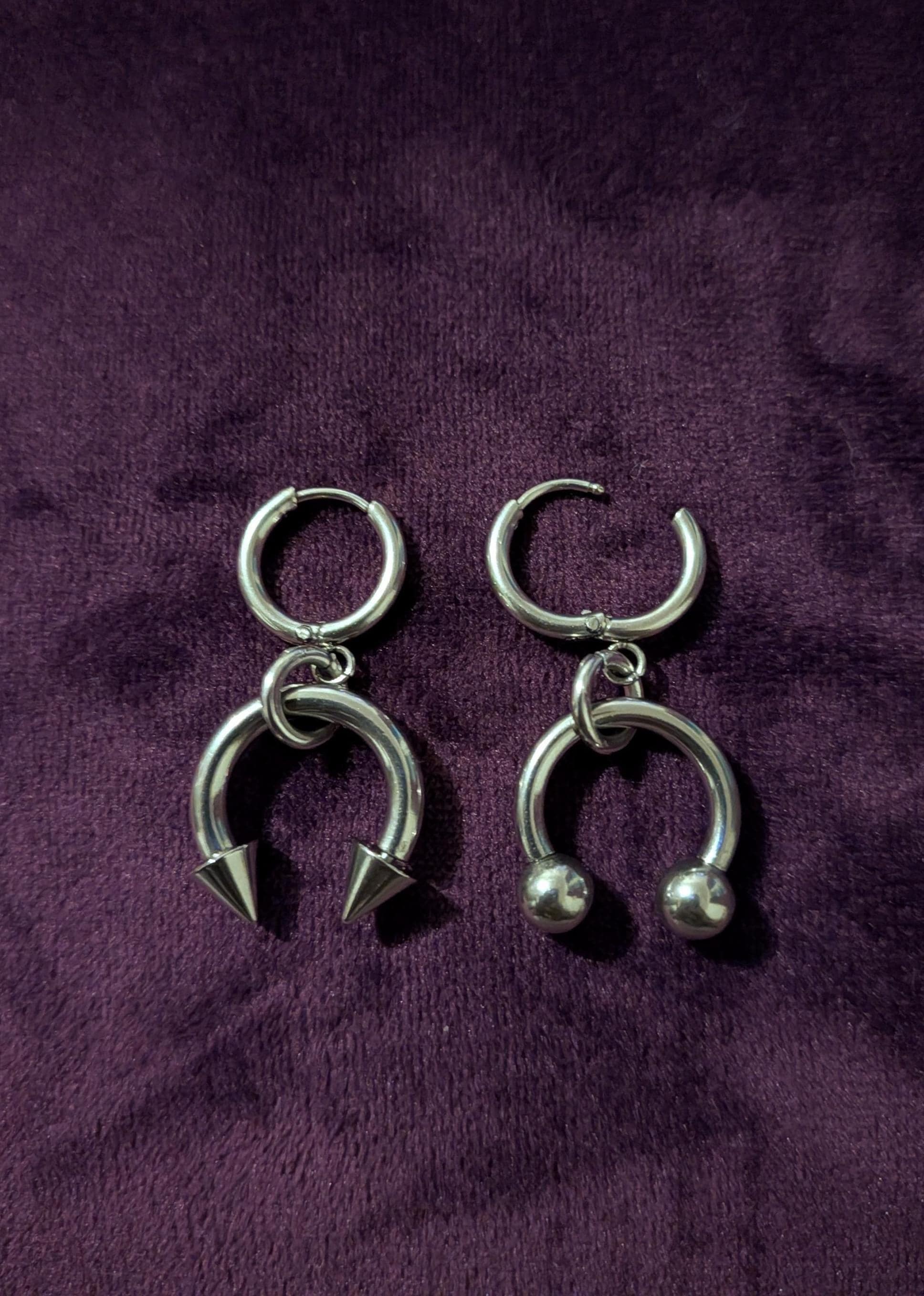 May include: A pair of silver-colored earrings. One earring features a hoop with a downward-facing, pointed spike. The other earring has a hoop with two small, round balls. Both earrings have a circular top.