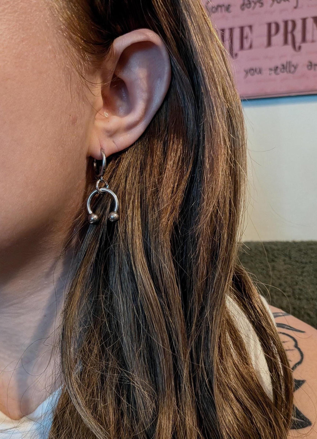 May include: Silver-toned horseshoe earrings with small ball accents. The earrings are attached to a small hoop that goes through the ear. The earrings are worn on a person's ear, with brown hair in the background. Jewelry.