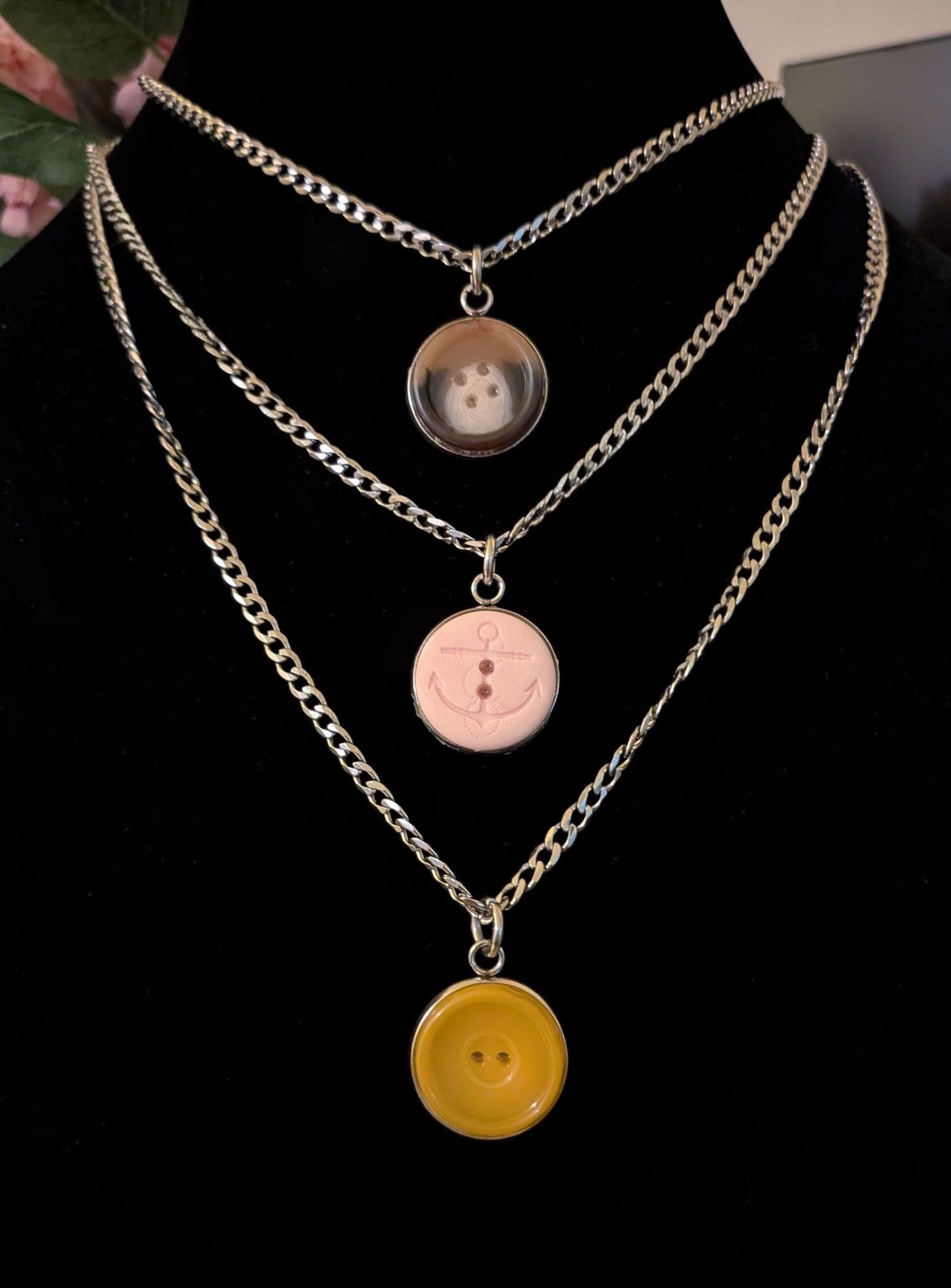 May include: Three-strand necklace set with silver-tone chains. Each chain features a different pendant: a brown button, a pink button with an anchor design, and a yellow button. The pendants are attached to the chains with silver-tone clasps.
