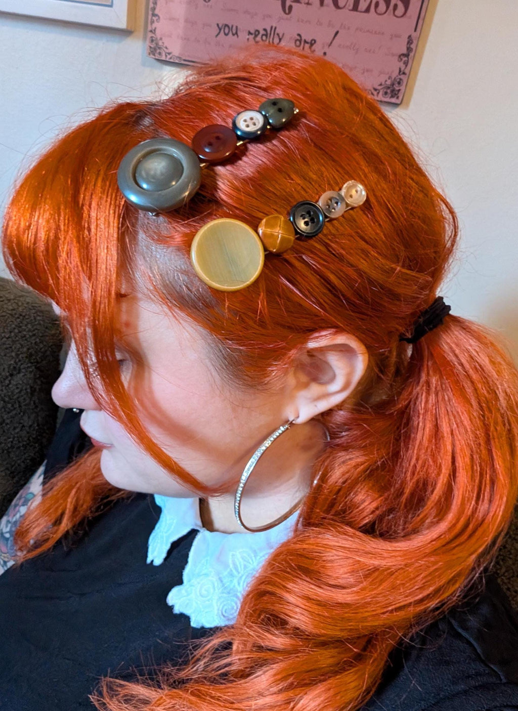 May include: A person with vibrant red hair styled in a ponytail, adorned with button hair clips in various colors. Large hoop earrings add a touch of sparkle. The hair accessories include buttons in shades of gray, brown, and yellow.