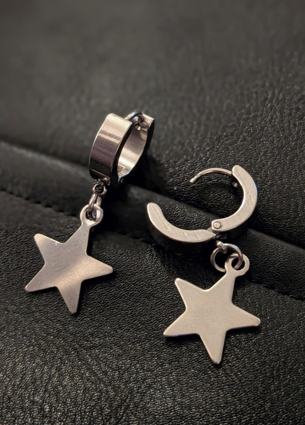 May include: A pair of silver-colored hoop earrings with a star-shaped charm. The earrings are made of a shiny metal and have a simple, modern design. The star charms hang from the hoops, adding a touch of whimsy.