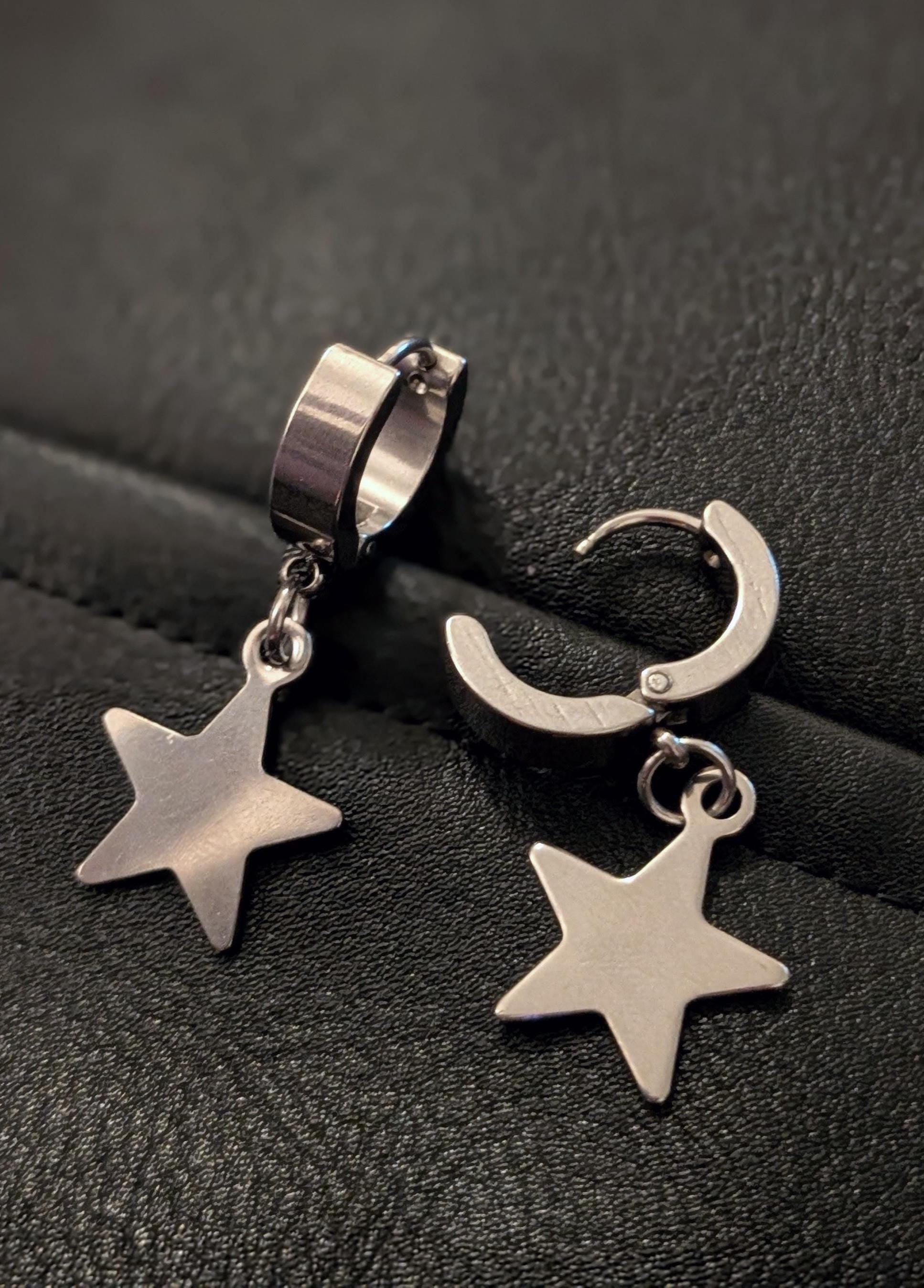 May include: A pair of silver-colored hoop earrings with a star-shaped charm. The earrings are made of a shiny metal and have a simple, modern design. The star charms hang from the hoops, adding a touch of whimsy.