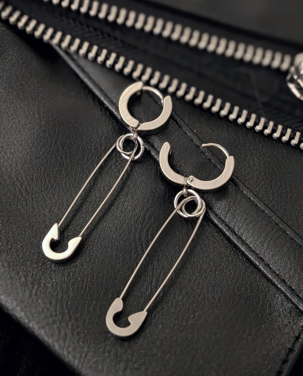 May include: Silver-toned hoop earrings with a safety pin design. The earrings are displayed on a black leather surface with a zipper detail in the background. The safety pins are attached to the hoops with small rings.