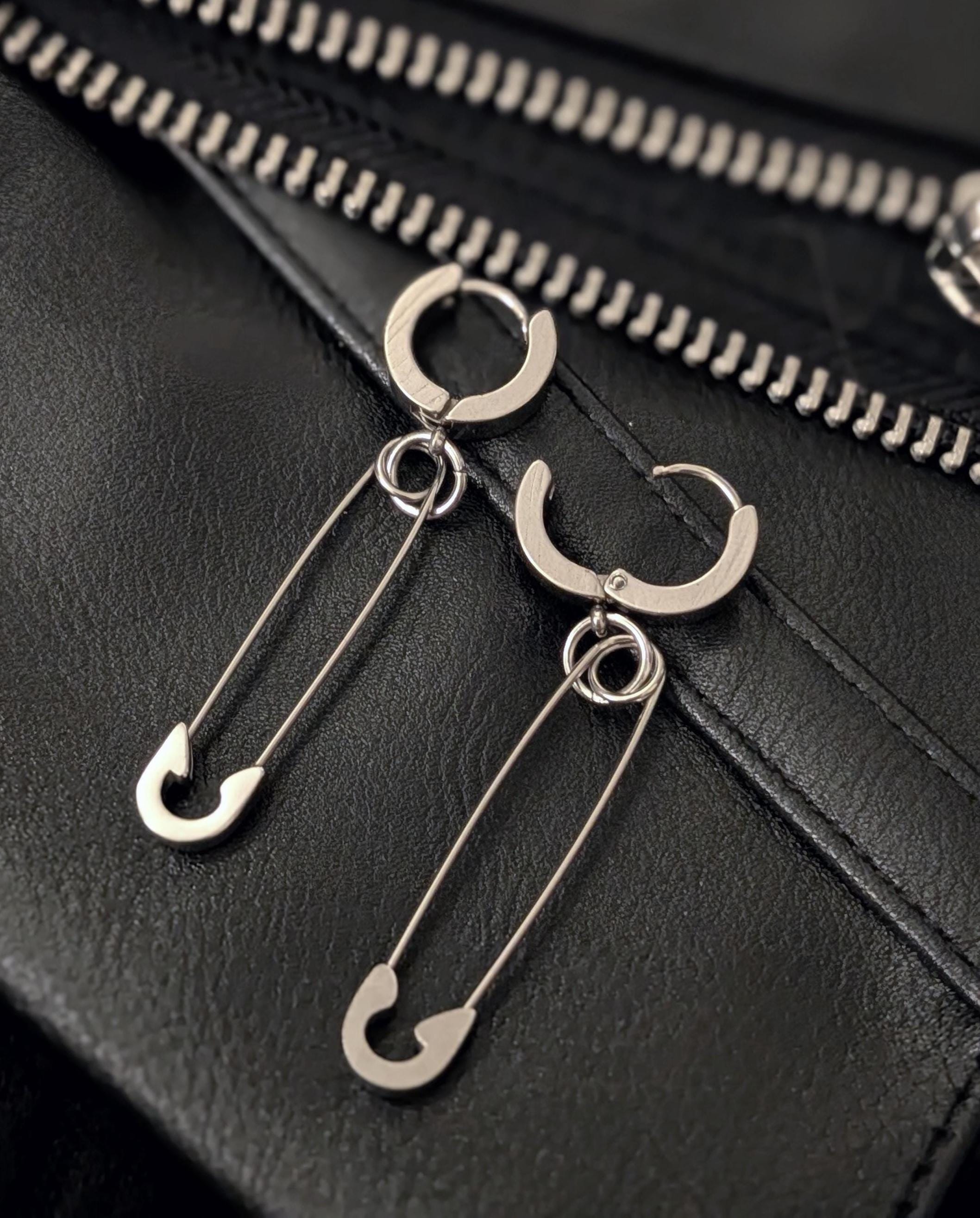 May include: Silver-toned hoop earrings with a safety pin design. The earrings are displayed on a black leather surface with a zipper detail in the background. The safety pins are attached to the hoops with small rings.