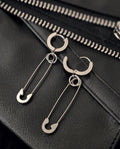May include: Silver-toned hoop earrings with a safety pin design. The earrings are displayed on a black leather surface with a zipper detail in the background. The safety pins are attached to the hoops with small rings.