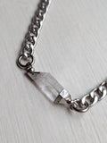May include: A silver chain necklace with a clear quartz crystal pendant. The crystal is rectangular and faceted, and the chain has large, interlocking links. The necklace is displayed on a white wooden surface.