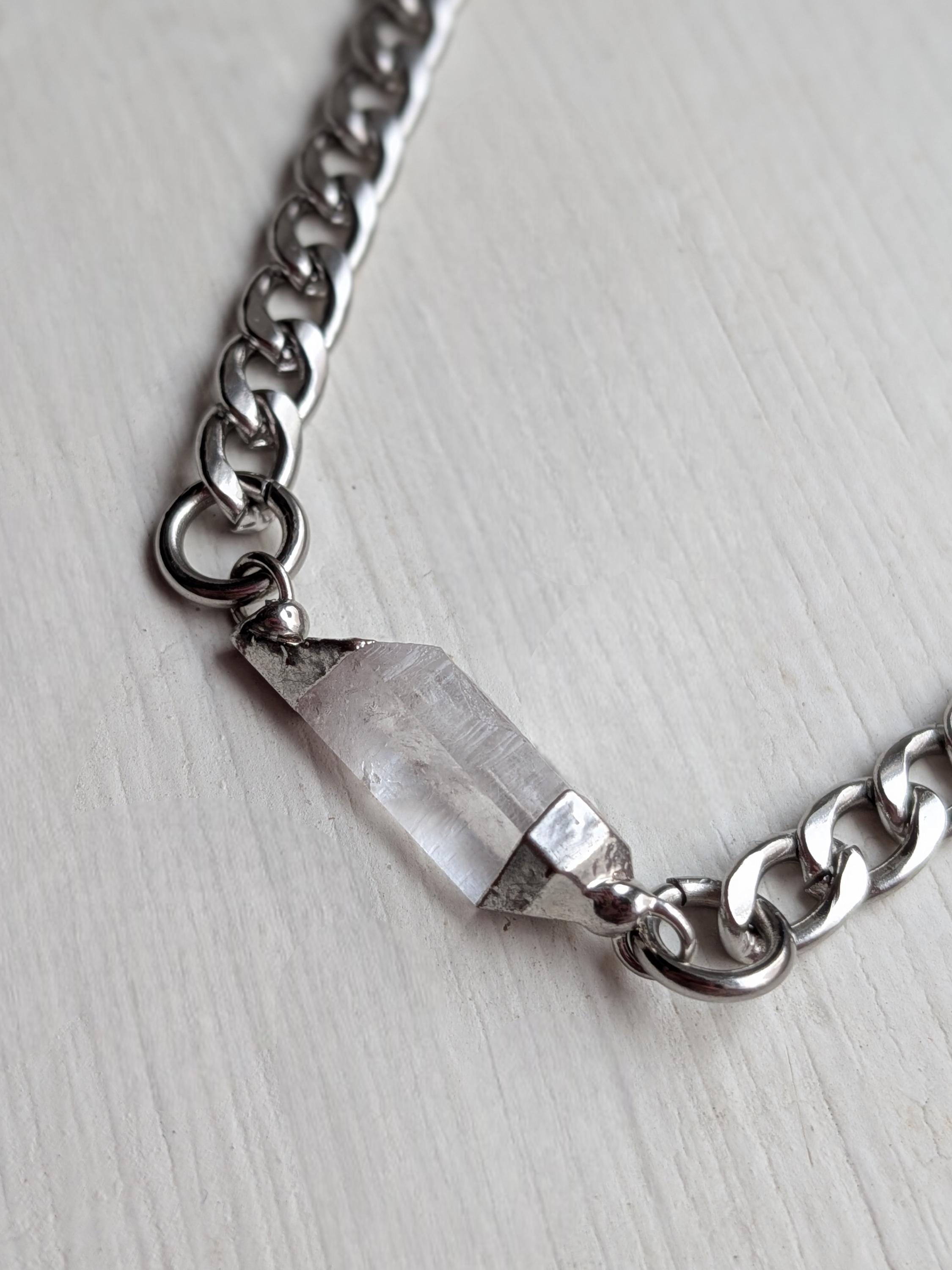 May include: A silver chain necklace with a clear quartz crystal pendant. The crystal is rectangular and faceted, and the chain has large, interlocking links. The necklace is displayed on a white wooden surface.