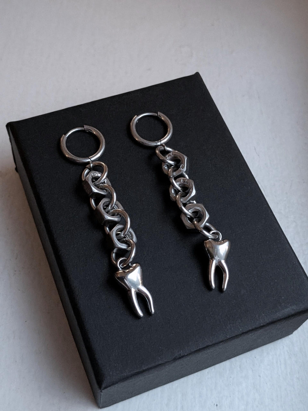 May include: A pair of silver-toned earrings. Each earring features a small hoop, connected to a chain of metal links, and a tooth charm. The earrings are displayed on a black box.