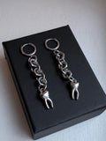 May include: A pair of silver-toned earrings. Each earring features a small hoop, connected to a chain of metal links, and a tooth charm. The earrings are displayed on a black box.