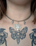 May include: Silver-toned necklace with a chunky chain. The pendant features a moth design with a sword and crown, and two tooth charms. The necklace is worn on a person's chest, with a large moth tattoo visible.