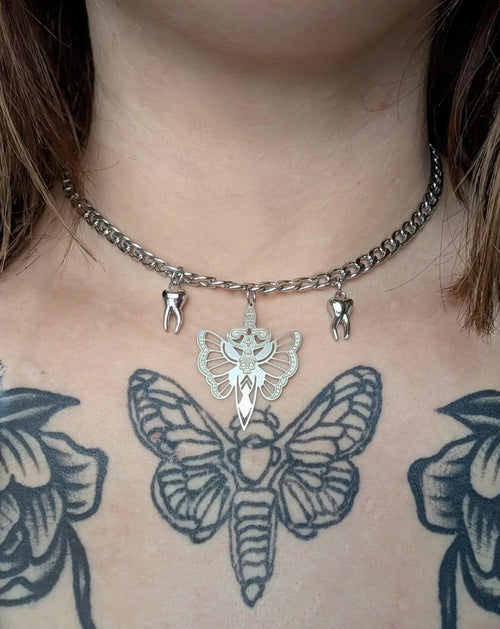 May include: Silver-toned necklace with a chunky chain. The pendant features a moth design with a sword and crown, and two tooth charms. The necklace is worn on a person's chest, with a large moth tattoo visible.