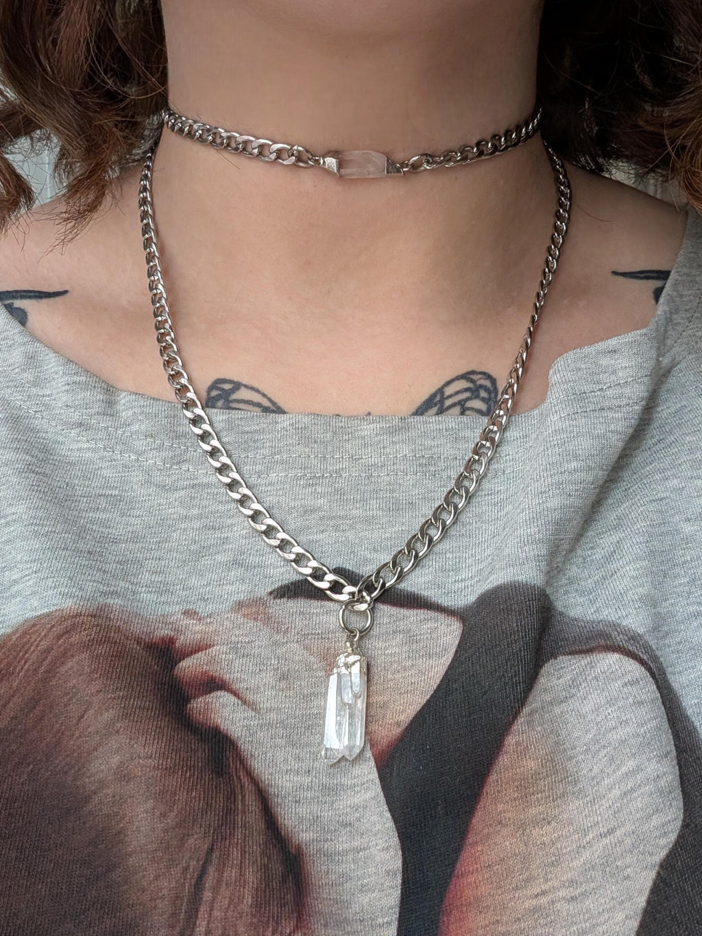 May include: A silver chain necklace with a clear crystal pendant and a matching choker. The choker features a rectangular pink stone. The necklace is worn over a gray t-shirt.