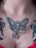May include: Silver-toned chain necklace with a large safety pin pendant. The safety pin holds three small tooth charms. The necklace is worn over a red lace top, with a black and gray butterfly tattoo visible.