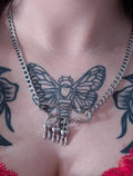 May include: Silver-toned chain necklace with a large safety pin pendant. The safety pin holds three small tooth charms. The necklace is worn over a red lace top, with a black and gray butterfly tattoo visible.