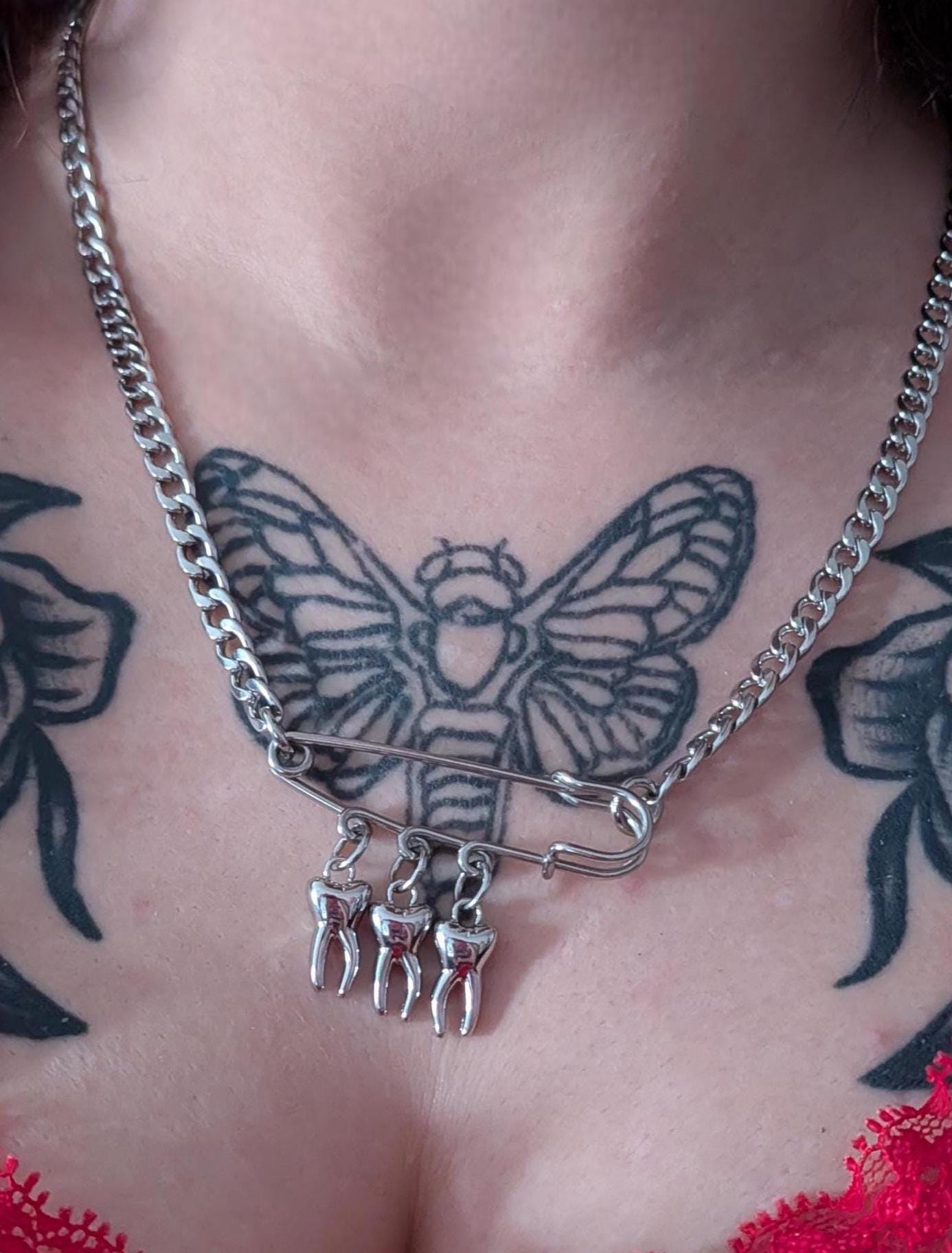 May include: Silver-toned chain necklace with a large safety pin pendant. The safety pin holds three small tooth charms. The necklace is worn over a red lace top, with a black and gray butterfly tattoo visible.