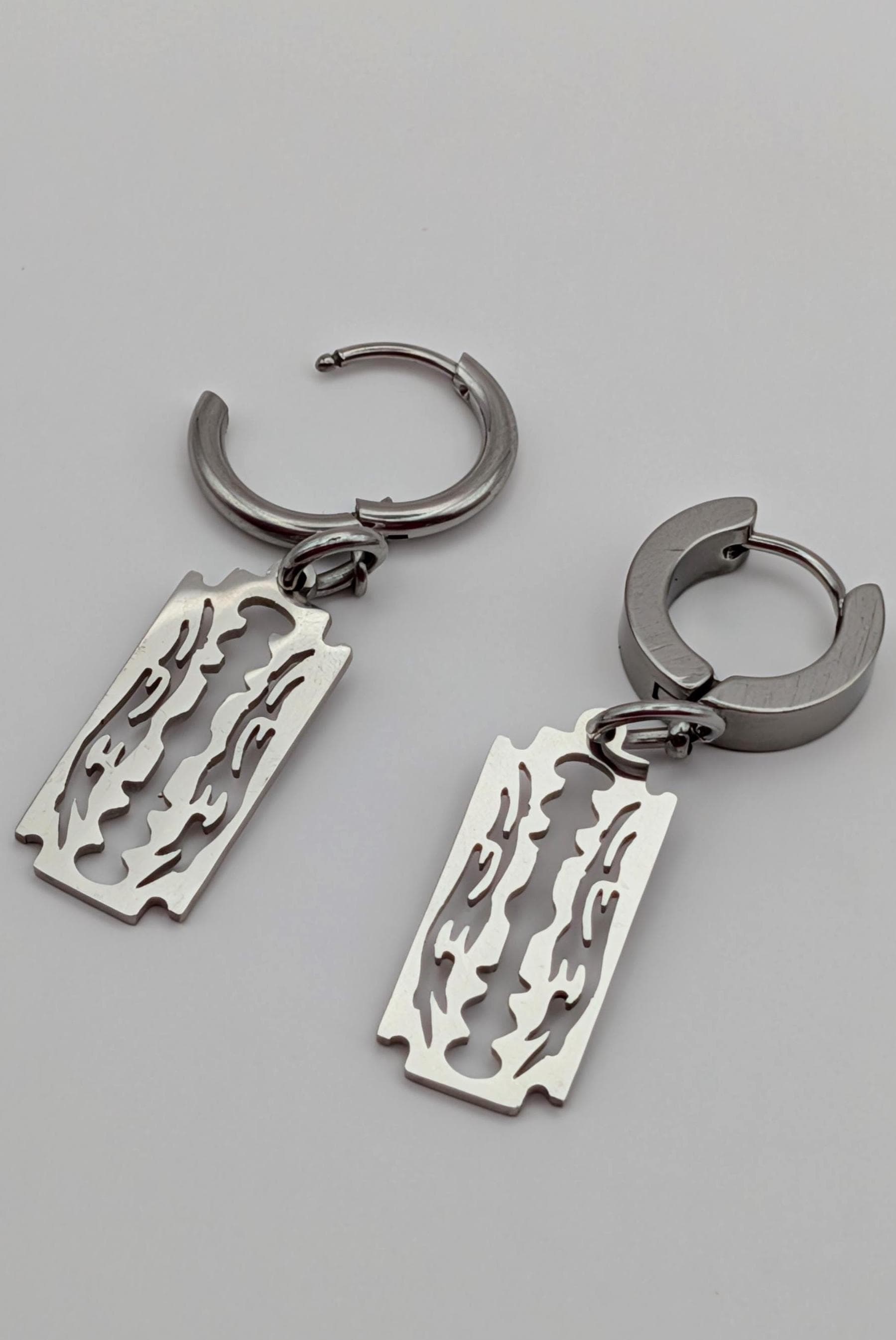 May include: A pair of silver-colored hoop earrings. Each earring features a rectangular charm with a decorative flame-like design. The earrings are shown against a light gray background.