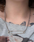 May include: A silver-colored necklace featuring a large safety pin pendant. The necklace has a chain link design and is worn around the neck. The safety pin is the focal point, adding a unique and edgy touch to the jewelry.